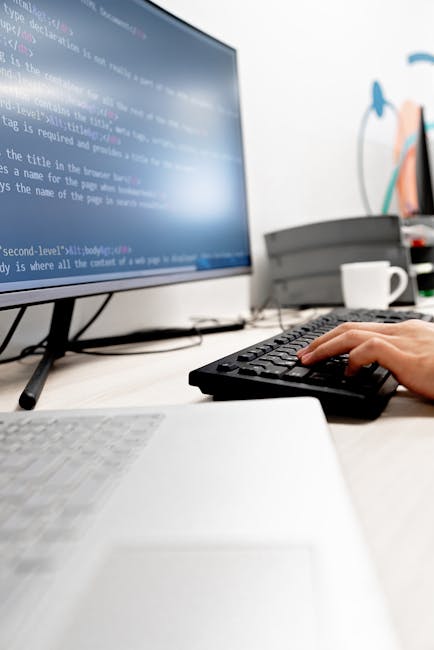 A close-up view of a computer monitor displaying lines of coding, with a person's hand operating a black keyboard in a modern office setting. The workspace includes a white desk with a white laptop pa